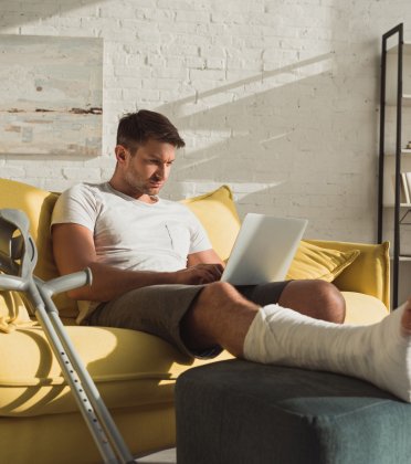 Een man heeft zijn been gebroken, rust uit in de zetel terwijl hij op de computer bezig is.